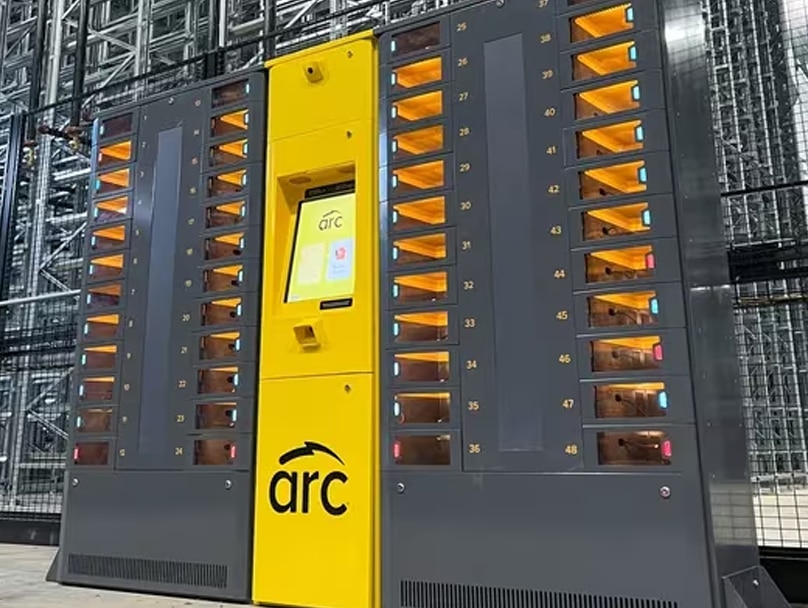 An ARC smart locker for device management with a yellow central panel displaying an "arc" logo and a yellow touchscreen interface. The machine has numerous numbered storage compartments on both sides, each illuminated in orange. The background shows a modern industrial setting.