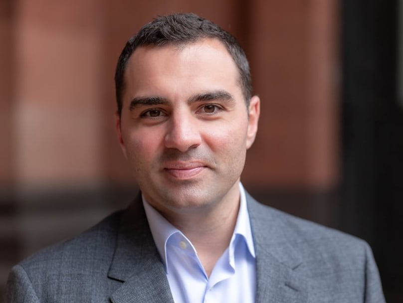 A man with short dark hair is smiling slightly, looking directly at the camera. He is wearing a light blue dress shirt and a grey suit jacket. The background is blurred, featuring tones of brown and black.
