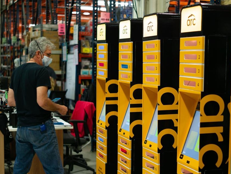 A man wearing a mask and glasses operates an ARC smart locker system in an industrial setting. To his right, there are several yellow and black "arc" branded devices lined up side by side. Shelves stocked with boxes and various items are visible in the background.