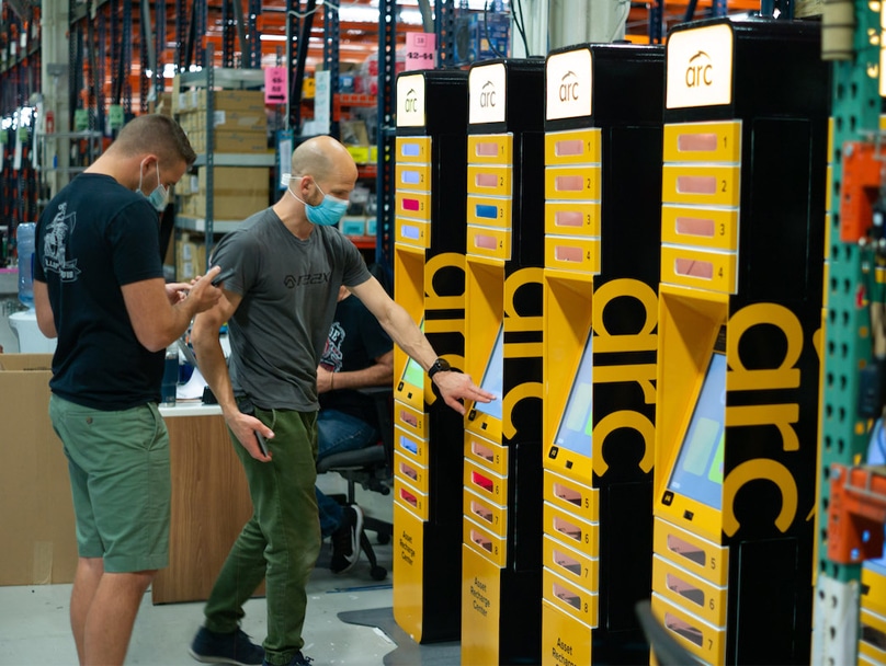 Two individuals wearing masks are interacting with a set of black and yellow ARC smart locker systems in a warehouse or industrial setting. Shelves filled with various items are visible in the background. One individual points at the screen while the other observes.
