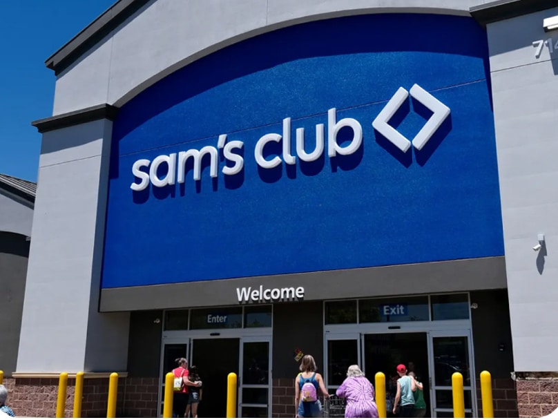 The entrance of a Sam's Club store, showing the large sign with the store’s name and logo above the doors. People are walking towards the entrance, and the words "Welcome," "Enter," and "Exit" are visible above the doors.