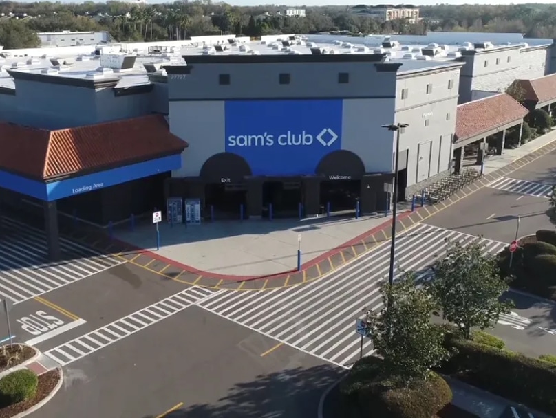 Aerial view of a Sam's Club store with a large blue sign on the front reading "sam's club". The entrance area is visible with marked exits and entrances. The parking lot in front has several cars parked and is surrounded by greenery.
