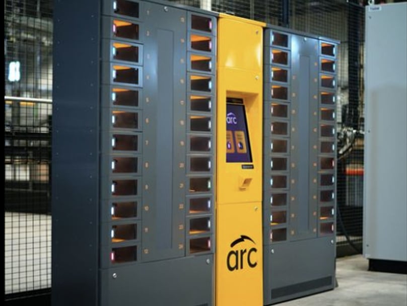 An ARC smart locker system with a yellow center panel labeled "arc" and multiple small compartments on both sides. The machine is placed indoors against a metal fence background. The compartments have small windows with lights inside.