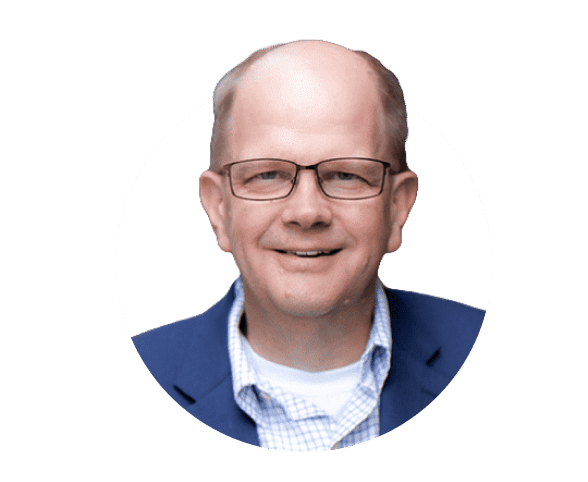 A man with short, light-colored hair and glasses is smiling. He is wearing a blue blazer over a checkered shirt. The background is plain white.