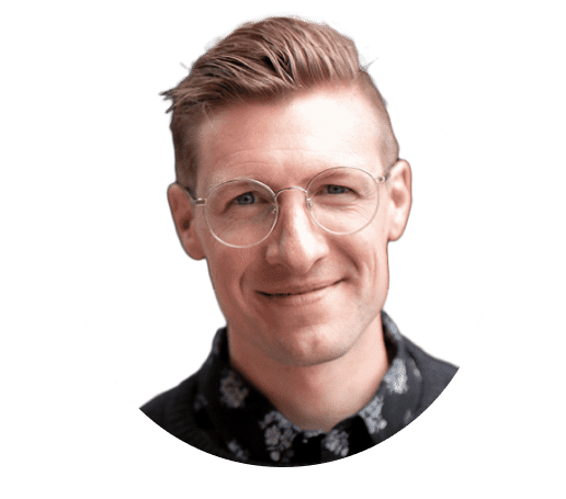 A person with short, light-colored hair and round glasses smiles at the camera. They are wearing a dark shirt with a floral pattern. The image has a circular frame and a white background.