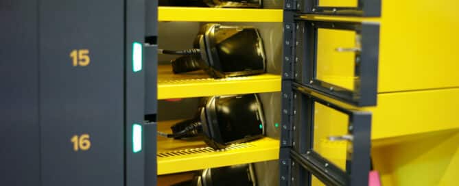 A close-up of an ARC intelligent locker system with multiple slots, numbered from 14 to 17. Each slot contains a mobile device being charged. The smart lockers have a black and yellow design, with blue lights visible on the left side indicating active charging.
