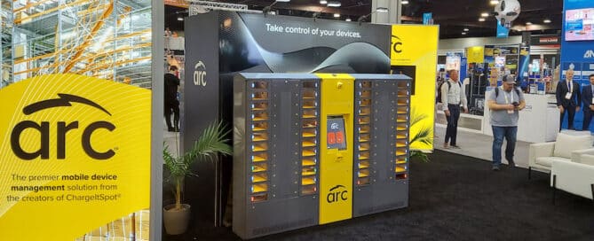 A trade show booth displays nn ARC intelligent locker. The booth, showcasing yellow and black branding, features intelligent lockers and a large sign that reads "Take control of your devices." Attendees are seen walking around the event space.