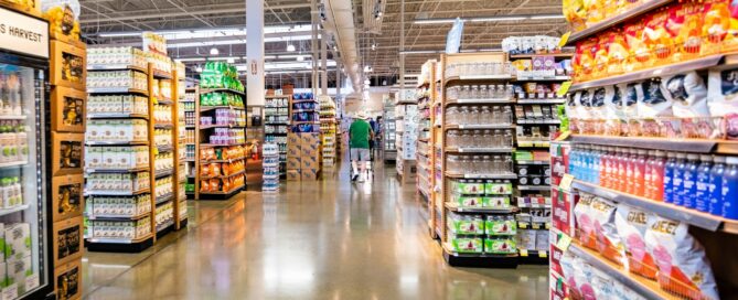 Wide aisle of a grocery store with various products on shelves, including snacks, drinks, and canned goods. The brightly lit, spacious area has a polished floor. A shopper in a green shirt is seen pushing a cart down the aisle.