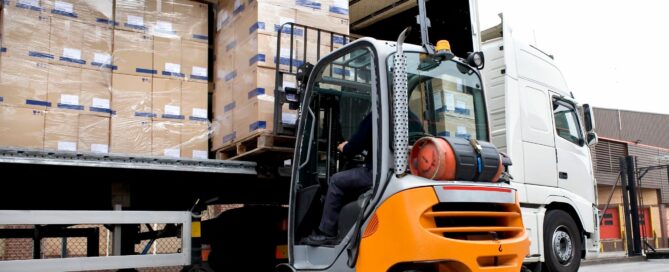 A forklift operator is unloading large pallets of cardboard boxes from the back of a semi-truck. The forklift is positioned on a concrete surface, while the truck's trailer door reveals the stacked boxes inside.