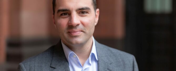 A person with short dark hair, wearing a light blue shirt and a grey suit jacket, stands in front of a blurred background with red and black tones. The individual is looking directly at the camera with a neutral expression.