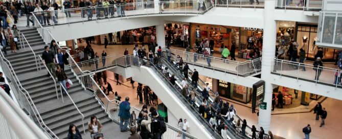 A busy multi-level shopping mall featuring numerous stores, an escalator, and a staircase. Shoppers are walking through the mall, browsing, and using the escalator and stairs. The mall has wide walkways and a modern design with tall glass windows and open spaces.