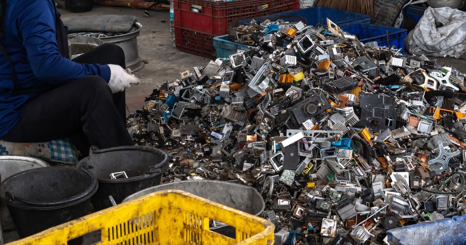 A person wearing a blue shirt and gloves is sitting next to a large pile of discarded electronic components and scrap metal. Buckets and crates surround the person, indicating the environmental impact of poor device management.