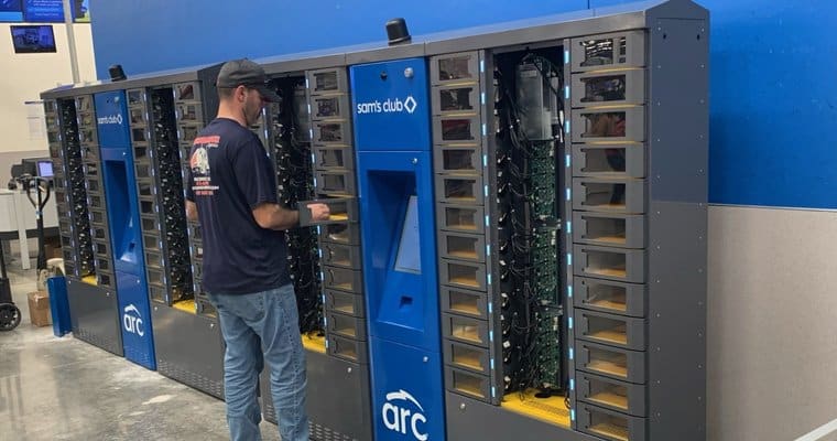 A man stands in front of a Sam's Club ARC SMART locker system, interacting with the touch screen. The Intelligent Lockers have multiple compartments and display the Sam's Club and ARC logos. The setting appears to be a well-lit indoor environment with various equipment visible.