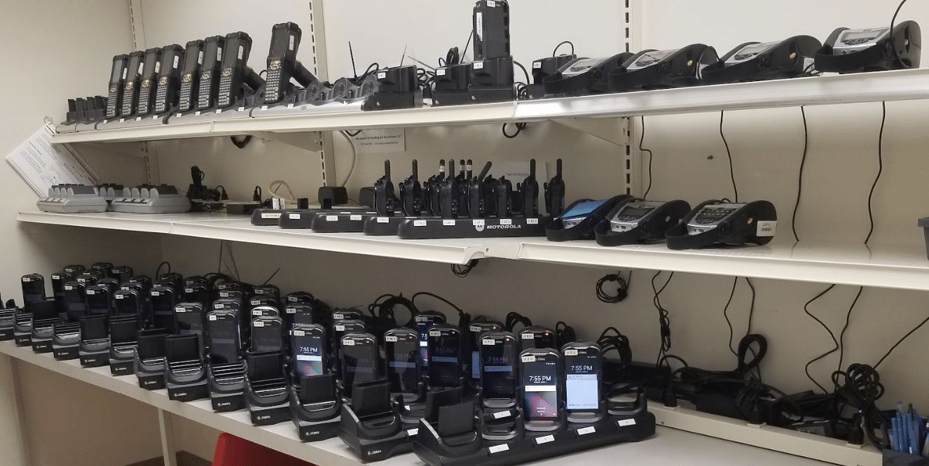 A warehouse worker in an orange safety vest and gray t-shirt is looking at a smartphone. The worker is standing in front of red metal shelving units filled with cardboard boxes and various items. The background shows a typical warehouse environment.