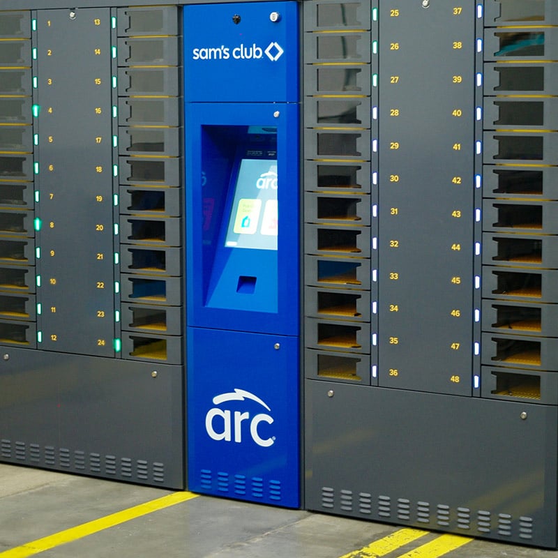 An ARC intelligent locker system branded for use by Sam's Club. The system has a blue central console with a touchscreen and numerous numbered lockers on both sides, for easy to use device management.The setup is placed on a warehouse floor with visible yellow safety lines.