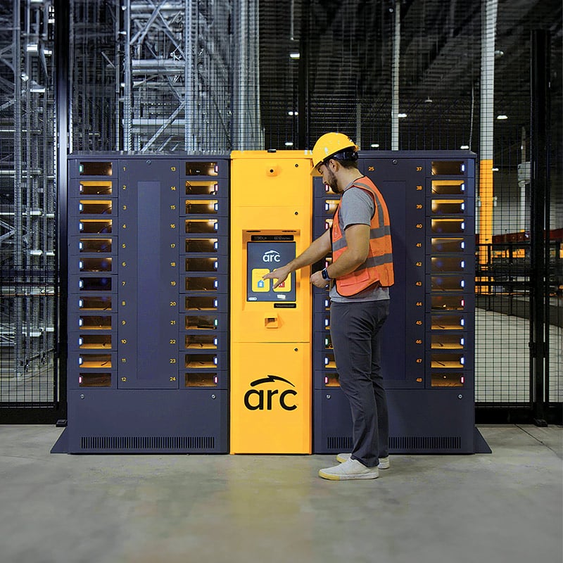 A worker in a safety helmet and vest operates an ARC smart locker in a warehouse. The unit has multiple compartments and a central touchscreen interface that the worker is interacting with.