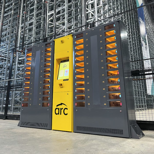 An ARC intelligent locker system with multiple compartments, each designated with a number, is located in a warehouse setting. The central yellow panel features a screen and the logo "arc." The surroundings include a high warehouse shelving system.