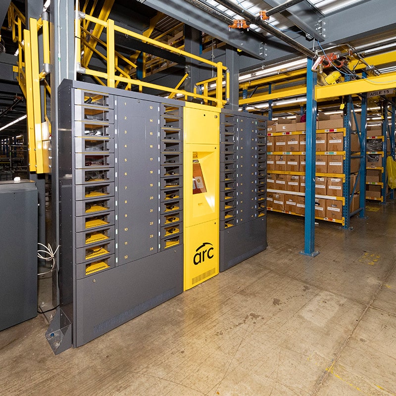 A large, automated storage and retrieval system sits in an industrial warehouse. The intelligent locker, labeled "arc," features multiple storage slots and a central control panel. Yellow structural elements and inventory shelves loaded with boxes are visible in the background.