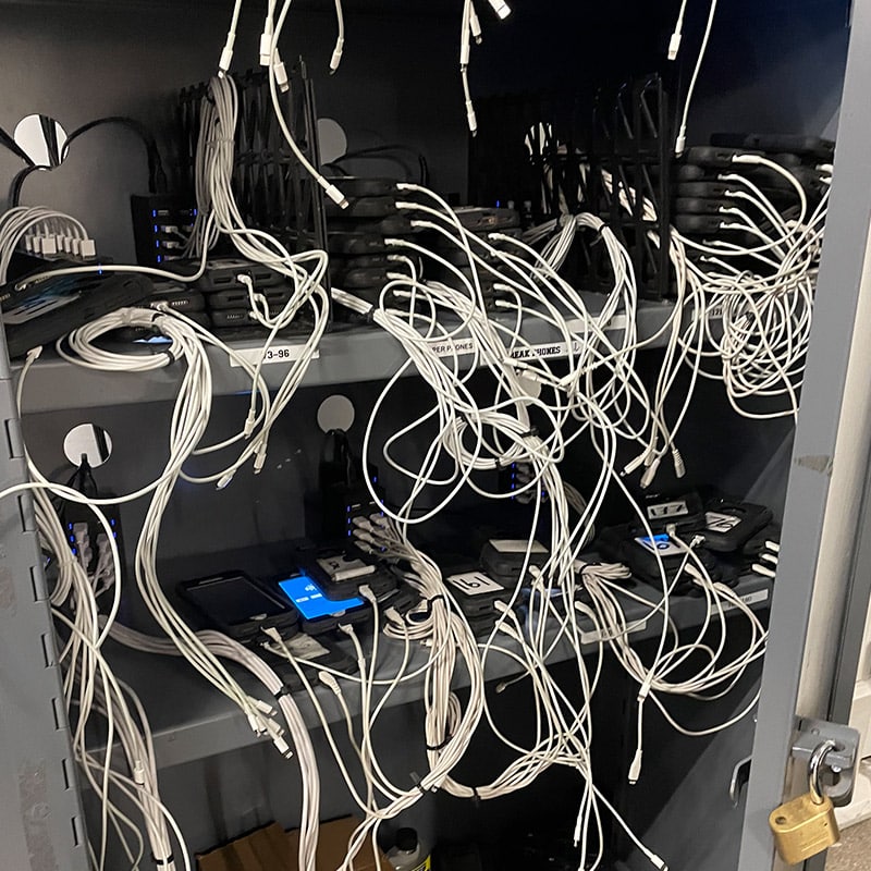 A cabinet filled with mobile devices, each connected with numerous white cables. The devices are labeled and arranged on multiple shelves. Some have blue indicator lights on, indicating they are charging.