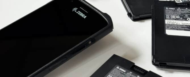 A black Zebra mobile device with a screen is placed on a white surface, surrounded by multiple rectangular black batteries or similar components. Some of the batteries are stacked on top of each other.