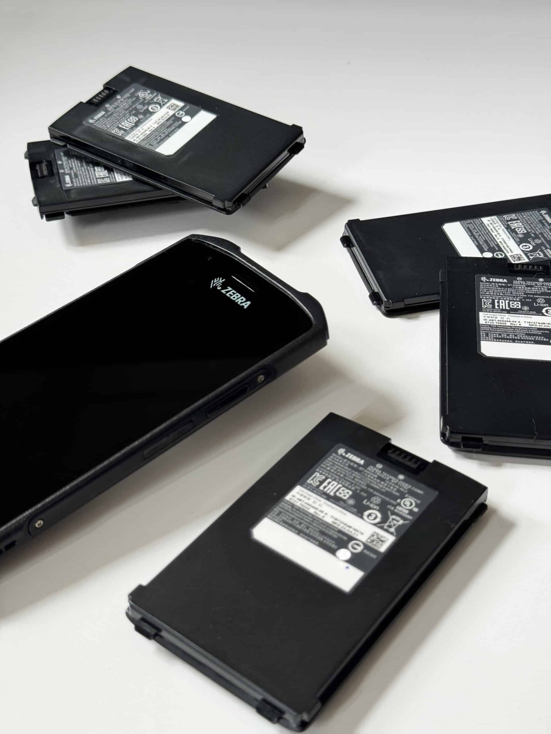 A black Zebra mobile device with a screen is placed on a white surface, surrounded by multiple rectangular black batteries or similar components. Some of the batteries are stacked on top of each other.