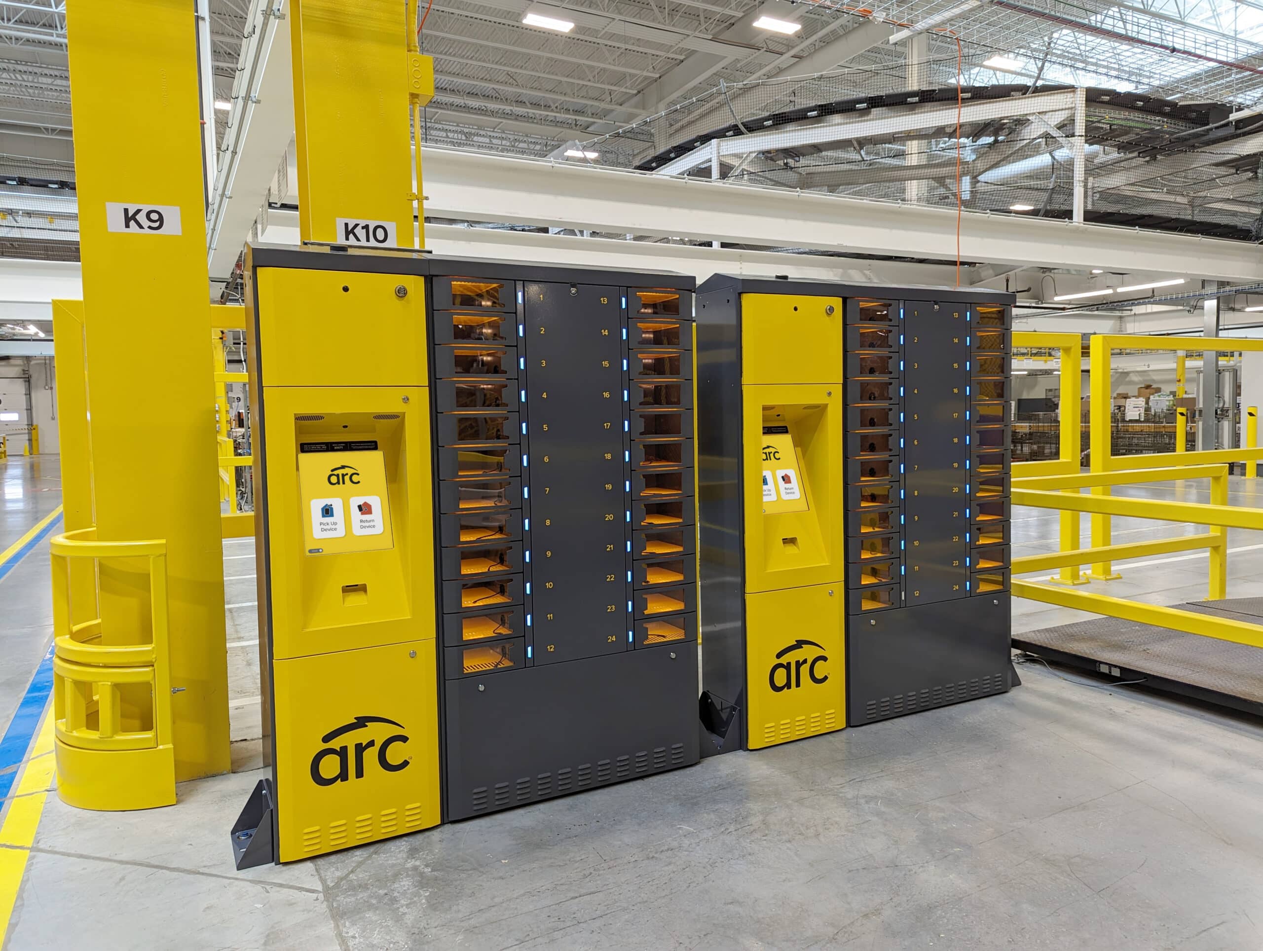 Two yellow and gray automated lockers with the label "arc" are positioned side by side in an industrial facility. Each locker features touchscreens for user interaction and multiple compartments for storage. The surrounding area includes structural supports and open space.