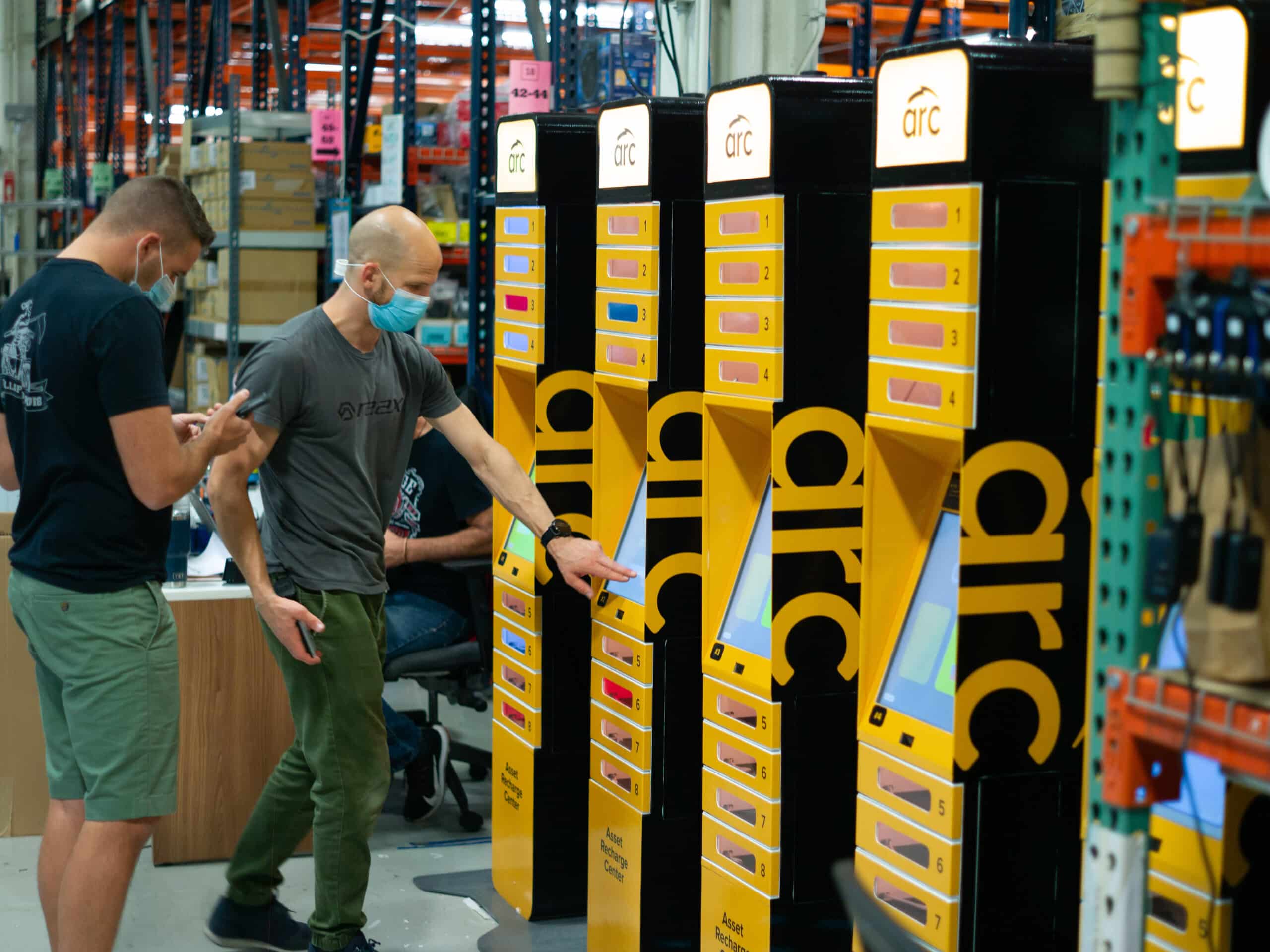 Two men inside a warehouse stand before a series of brightly lit yellow storage lockers labeled "arc." The man on the left checks his phone, while the man on the right, wearing a mask, interacts with one of the lockers, possibly retrieving an item.