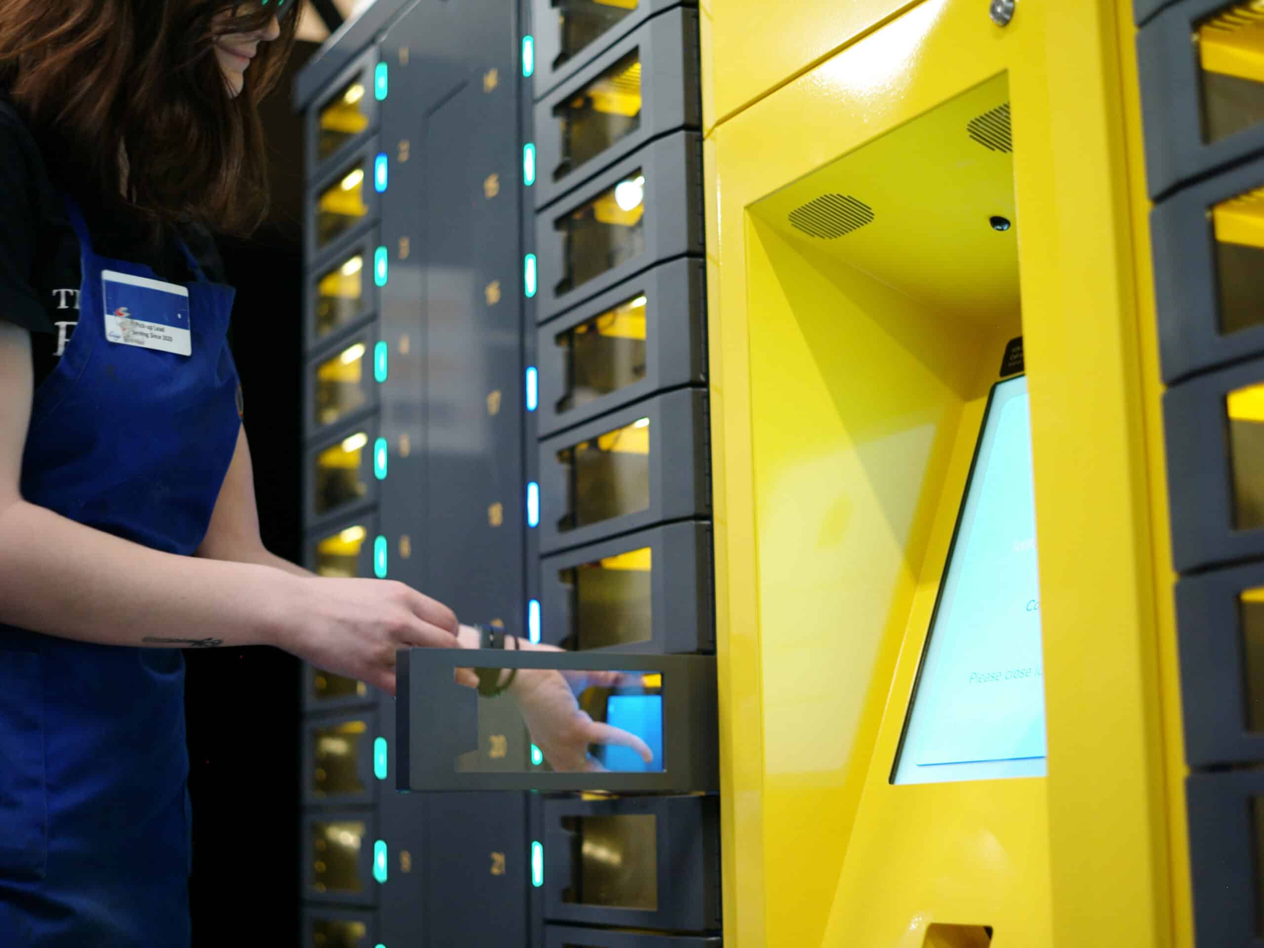 A person in a blue apron accessing a yellow, touchscreen locker system. The locker has multiple compartments, some open, with illuminated green lights indicating availability. The screen displays a message, and the person's hand is reaching into one compartment.