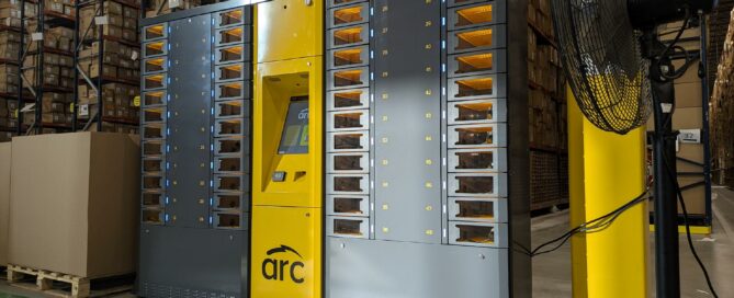 A large automated storage and retrieval system for employee devices stands in a warehouse. The unit has a yellow control panel in the center with the brand "arc" visible on it. Surrounding the control panel are multiple compartments filled with small containers.
