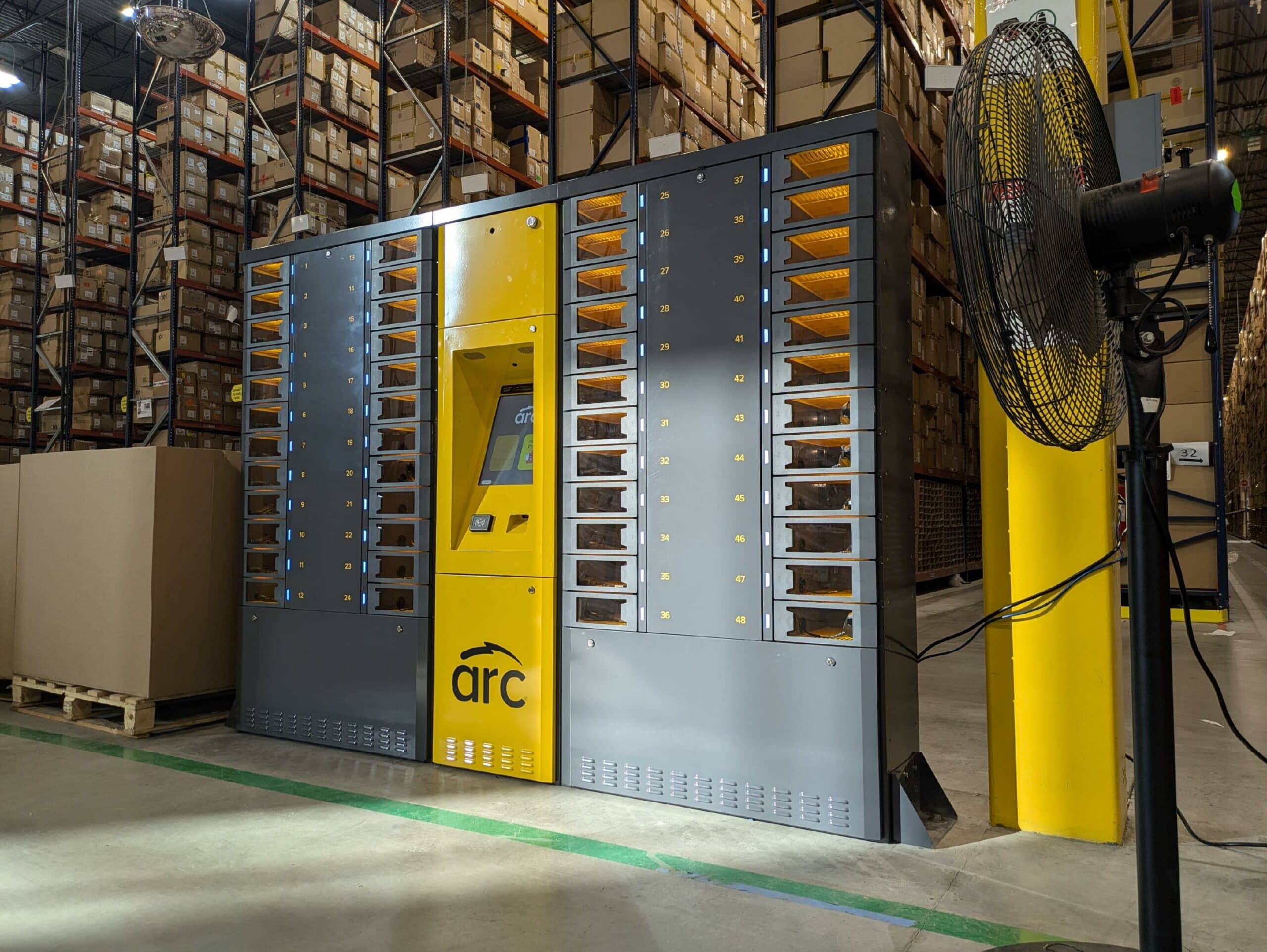A large automated storage and retrieval system for employee devices stands in a warehouse. The unit has a yellow control panel in the center with the brand "arc" visible on it. Surrounding the control panel are multiple compartments filled with small containers.