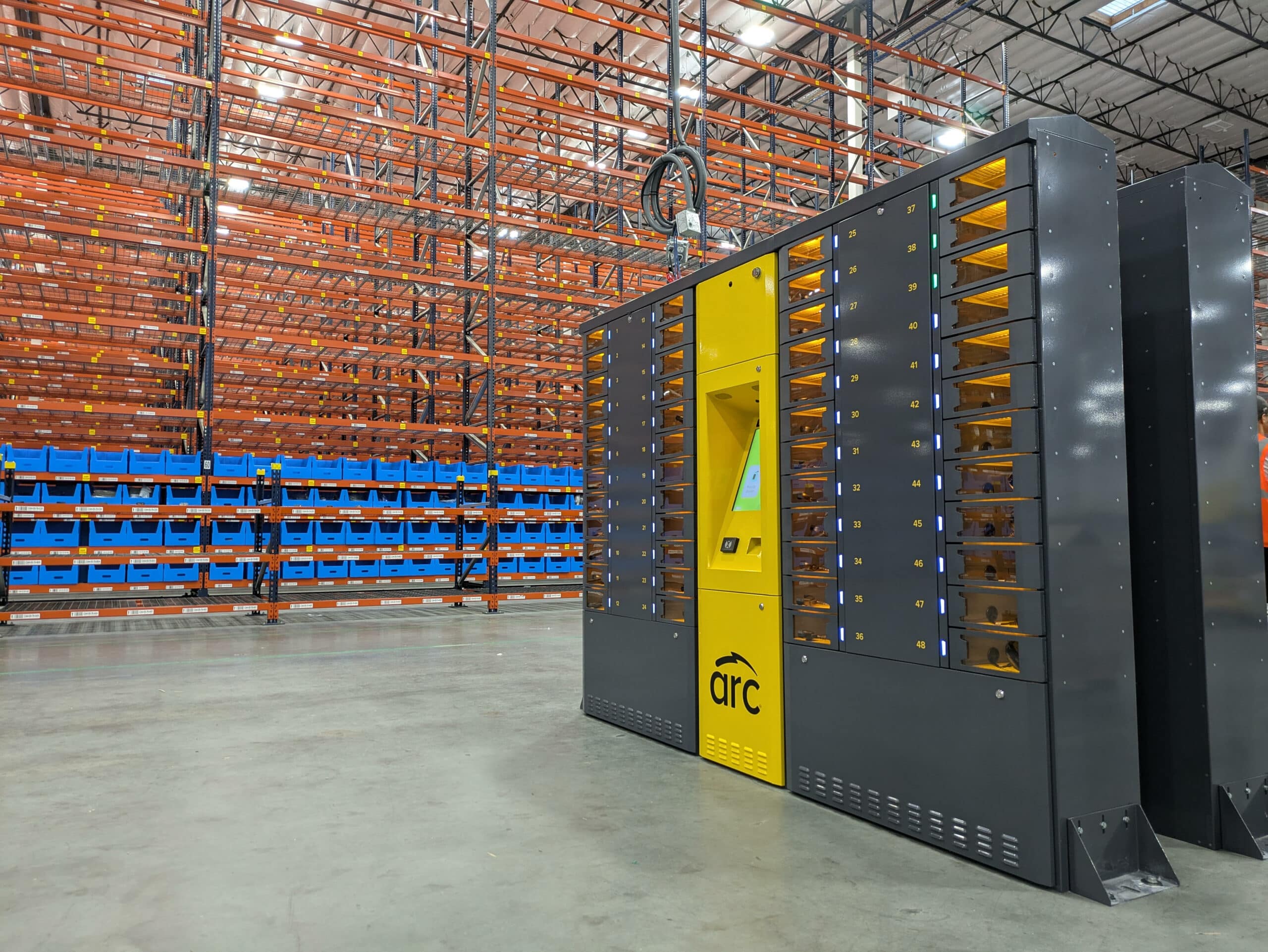 A large industrial warehouse with tall metal shelving units. In the foreground, there is a yellow and black automated robotics machine labeled "arc," featuring a screen interface. The shelves are mostly empty, aside from some blue bins.