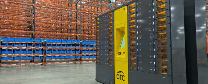 A large warehouse with tall racks filled with blue bins. In the foreground, a yellow and black automated machine with multiple compartments marked "arc" is positioned on the grey concrete floor. The high ceiling is supported by metal beams.