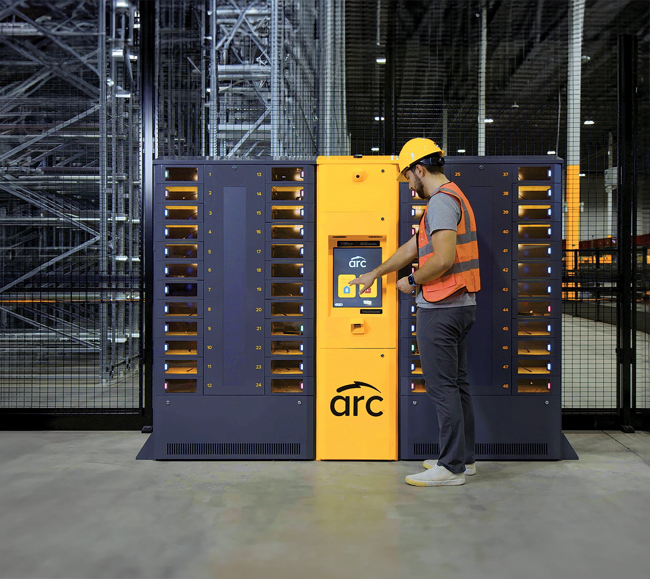 A worker in a hard hat and safety vest interacts with a yellow and black automated storage and retrieval system labeled "arc" in a warehouse setting. The system has multiple compartments and a digital interface screen.