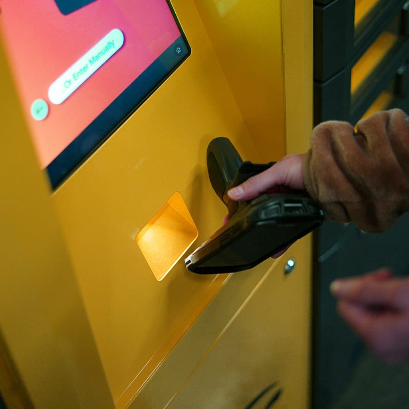 A person is scanning a QR code from a smartphone at an ARC intelligent locker system. The screen displays some text and an option to "Enter Manually." The person's hands are visible, holding the smartphone in place.